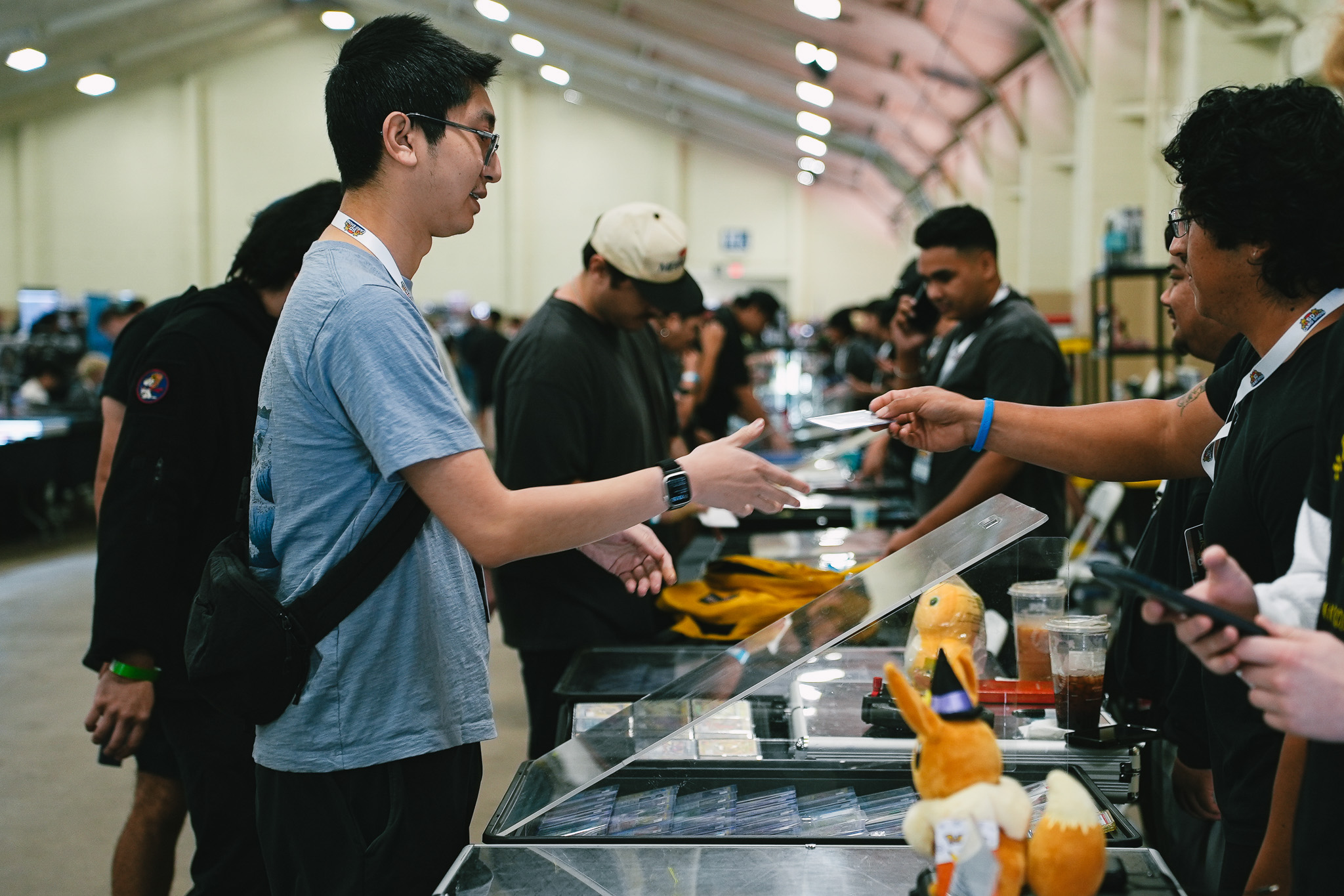 Two attendees exchanging a trading card at a vendor table, with Pikachu plush toys in the foreground